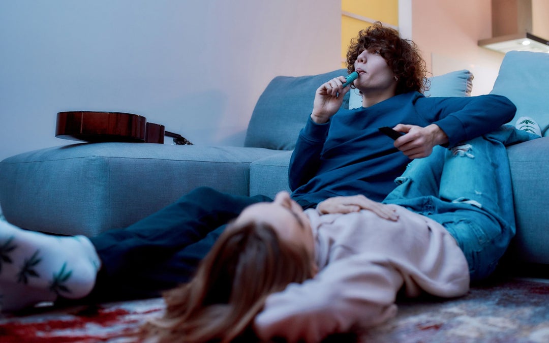 Young man smoking weed vape while lying on the sofa. Girlfriend is lying next him relaxing.