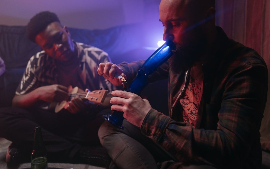 Two young African American men relaxing in a room. One of them is smoking weed from a bong while the other is playing on the guitar.