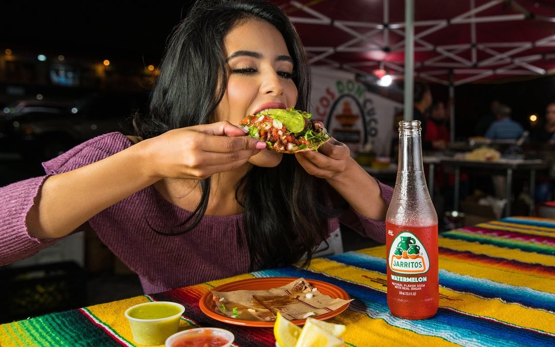 Hispanic lady has the munchies and is biting down into a huge soft-shell taco that she can hardly hold in her two hands.