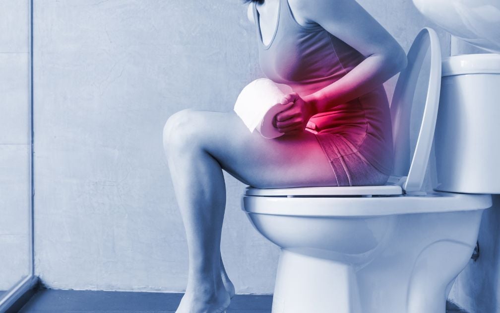 A young woman with digestive issues. She is sitting on a toilet bowl, holding her stomach. Her stomach is highlighted red to show her issues.
