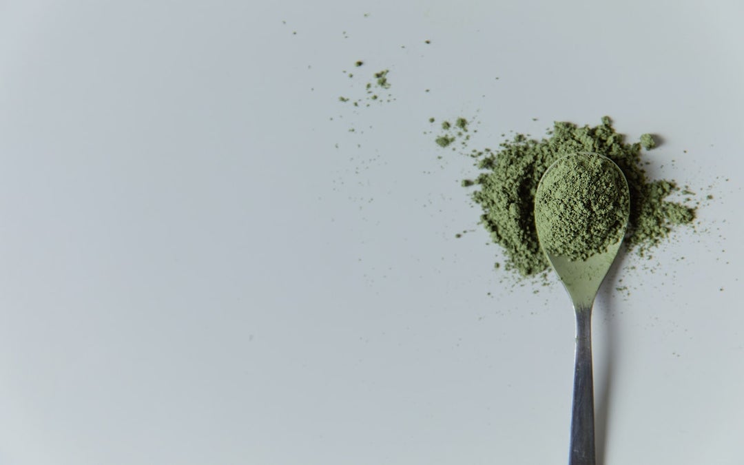 Green Kratom. Green-coloured powder sits on a silver spoon and white background.